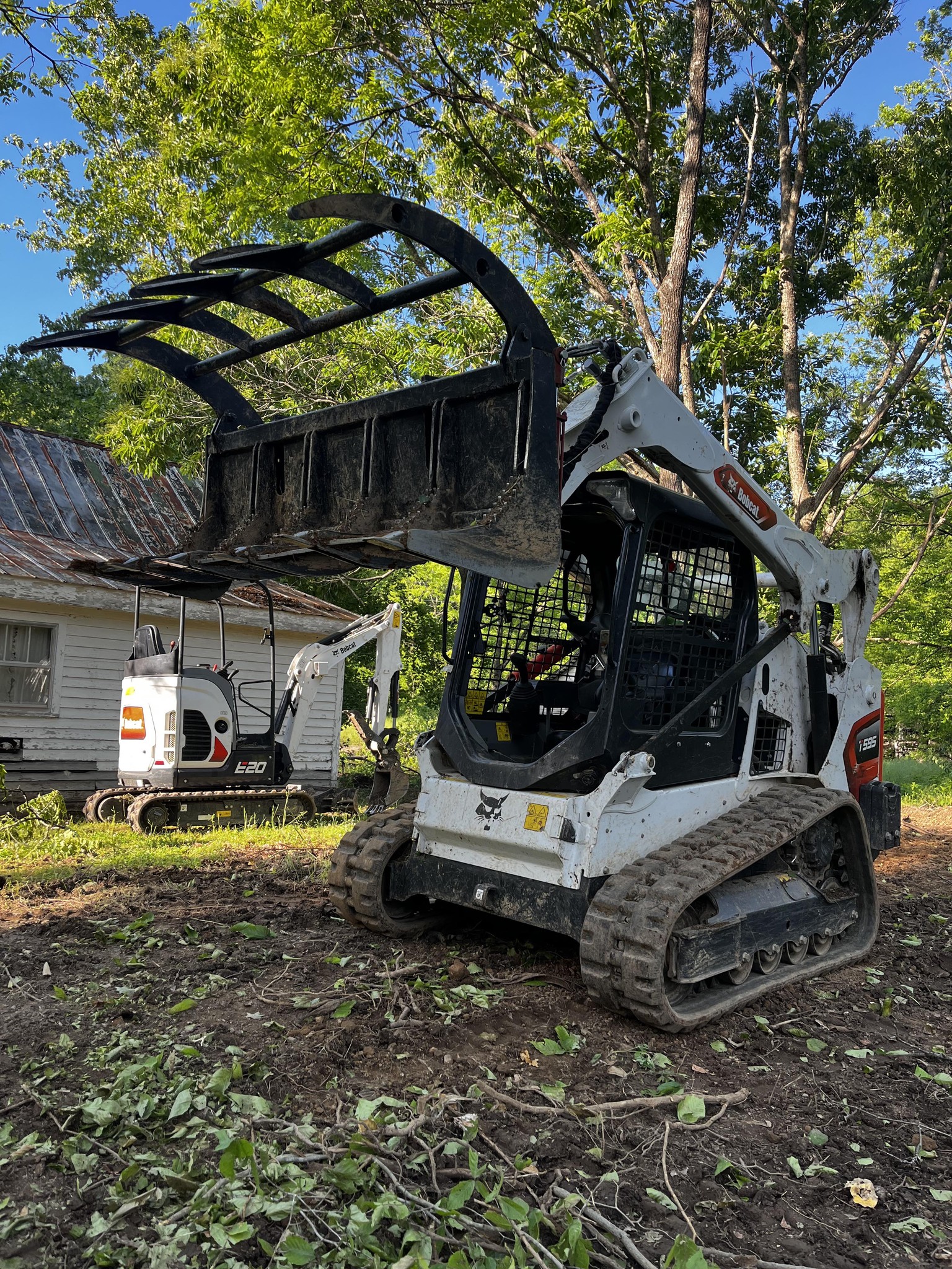 A white skid steer loader with a raised grapple attachment is parked on dirt near a house and another construction vehicle, surrounded by trees and foliage.