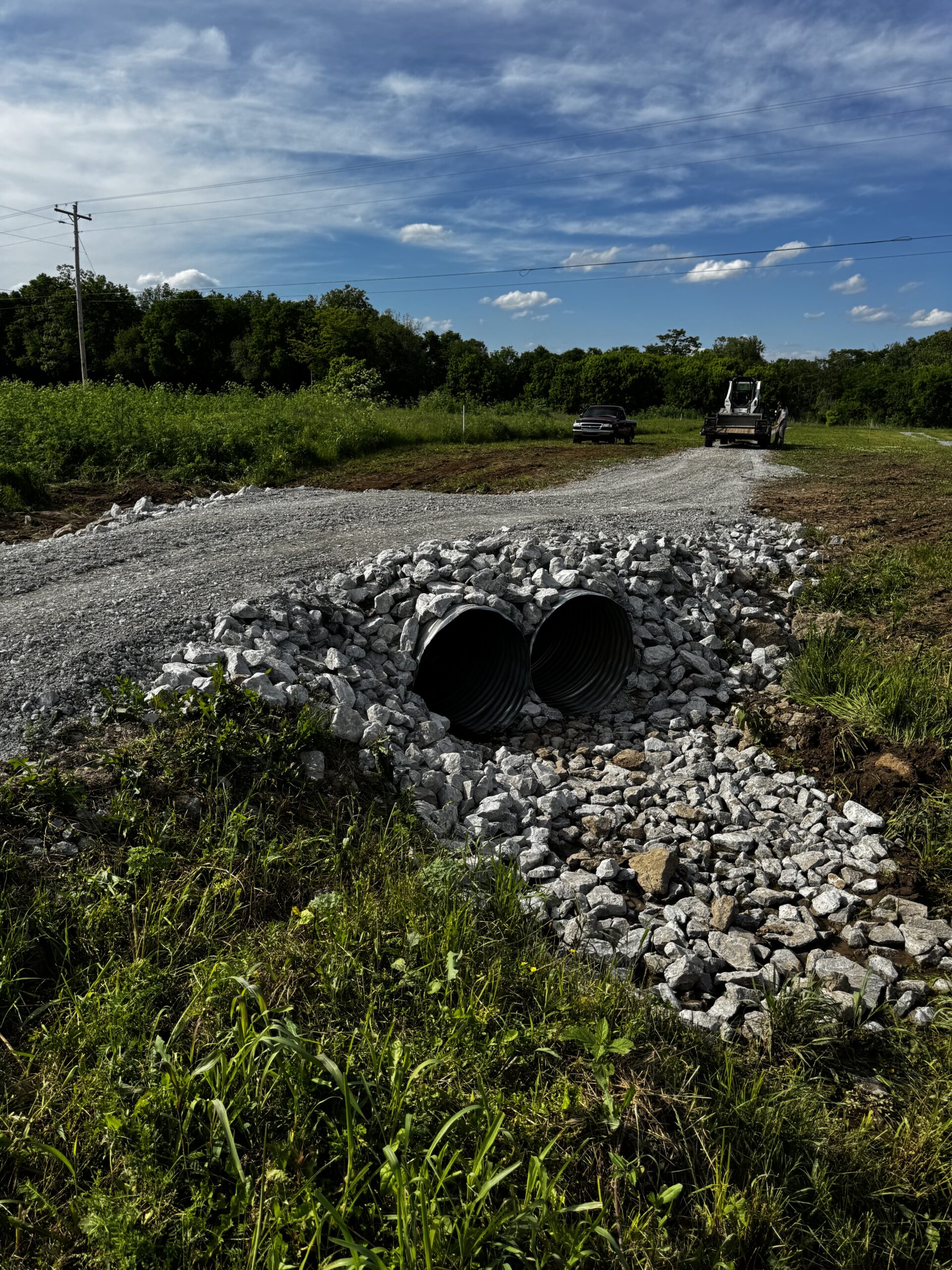 A gravel road under construction with two metal culvert pipes beneath it, surrounded by loose rocks and vegetation, with vehicles and trees in the background.