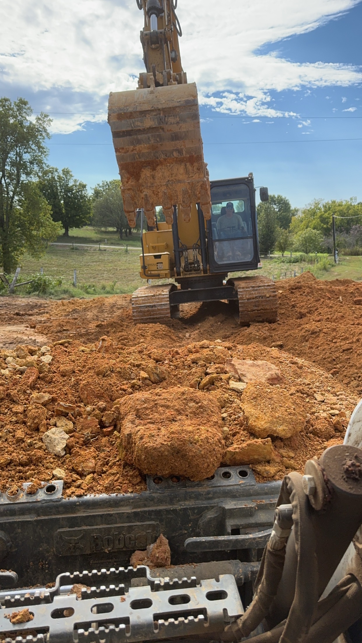 A yellow excavator scoops up reddish-brown dirt at a construction site with trees and a blue sky in the background.