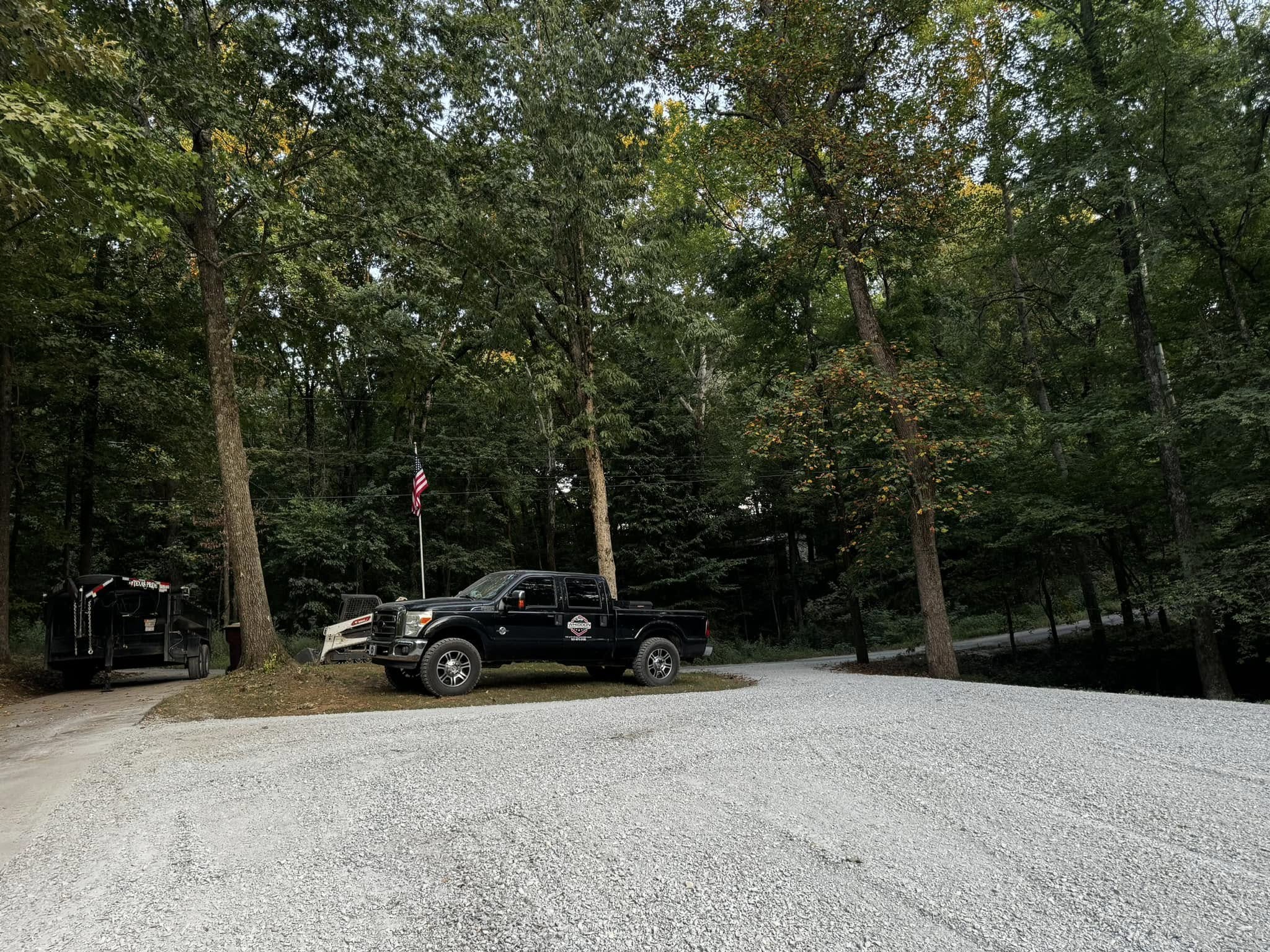 A black pickup truck and a trailer are parked on a gravel area surrounded by tall trees, with an American flag visible in the background.