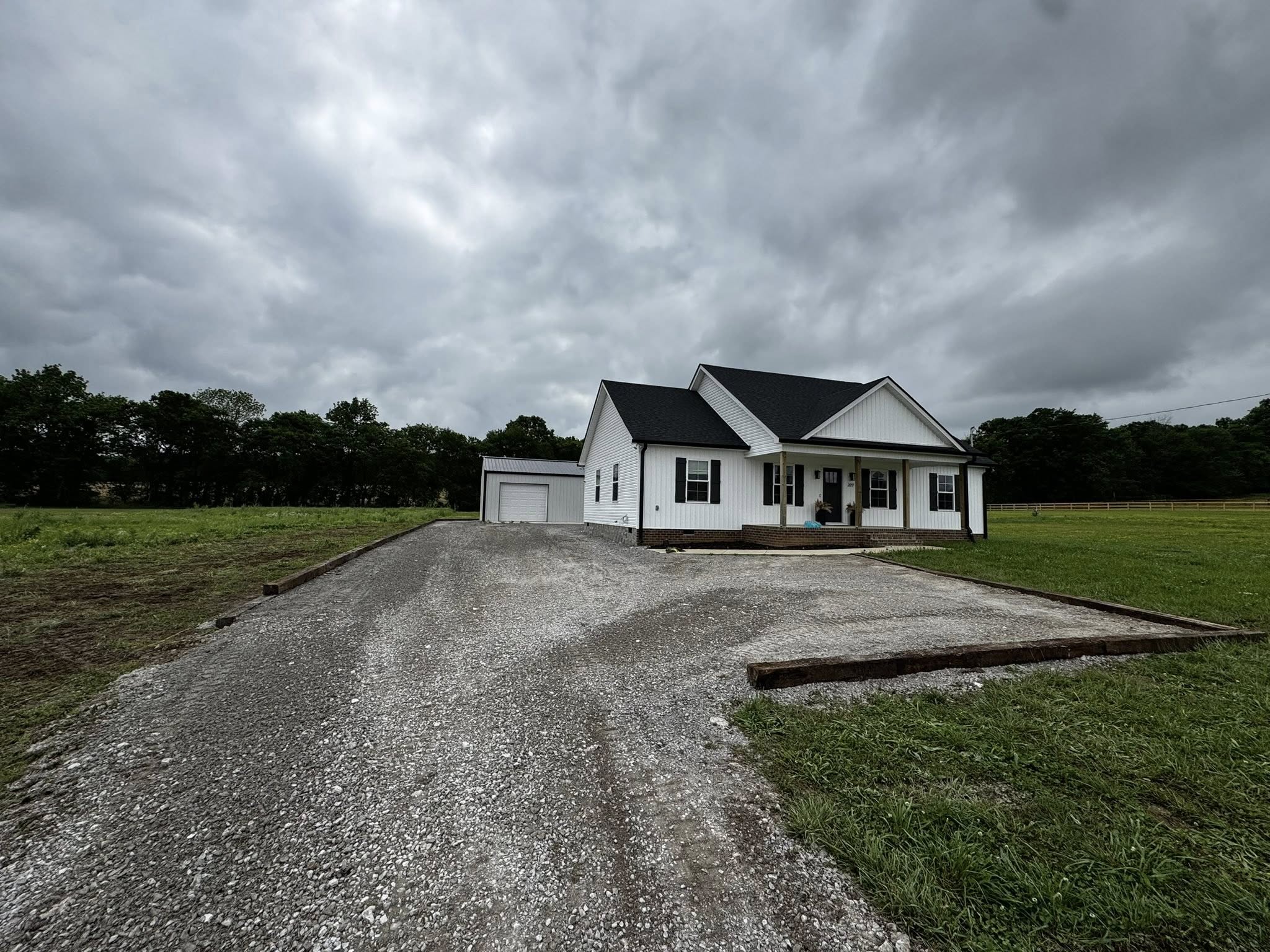 A white single-story house with a front porch and attached garage sits on a gravel driveway, surrounded by grass and trees under a cloudy sky.
