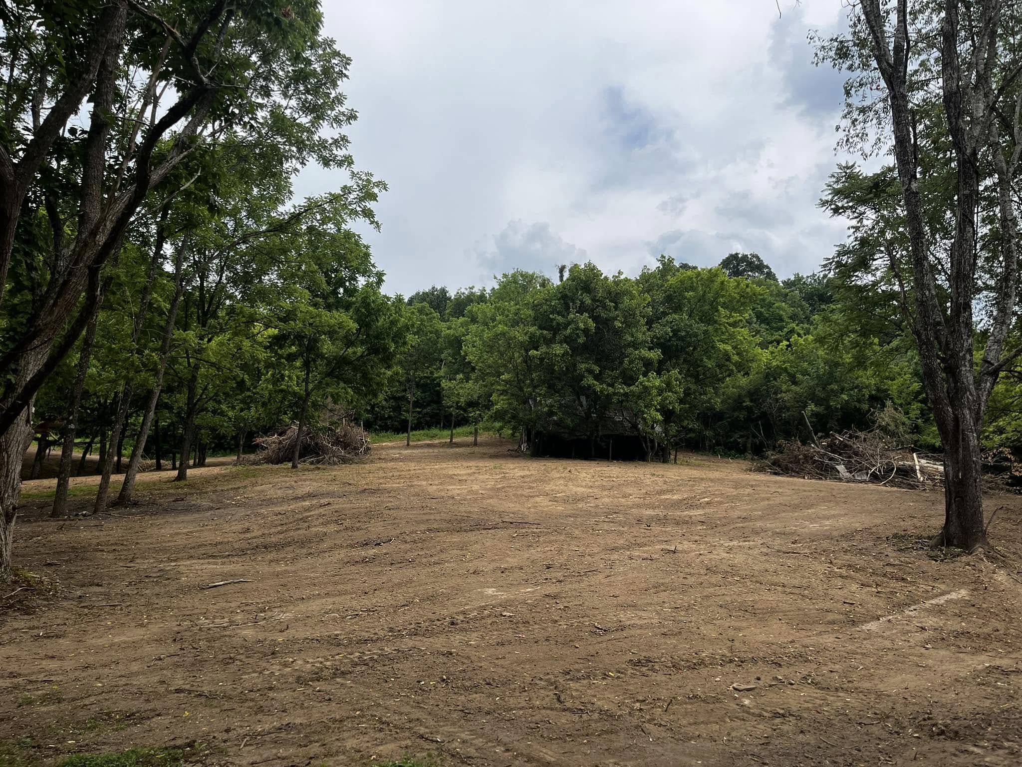 Cleared dirt area surrounded by trees and brush piles under a partly cloudy sky.