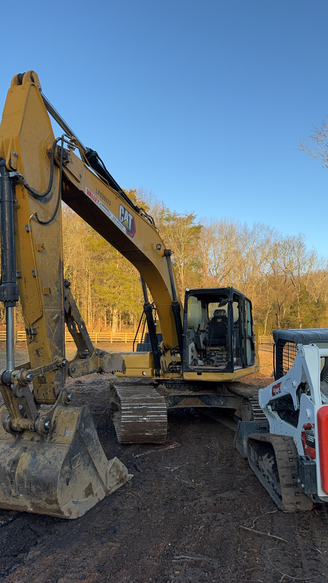 A yellow CAT excavator and a white Bobcat skid steer loader, ideal for excavation services, are parked on dirt at a construction site with trees and blue sky in the background.