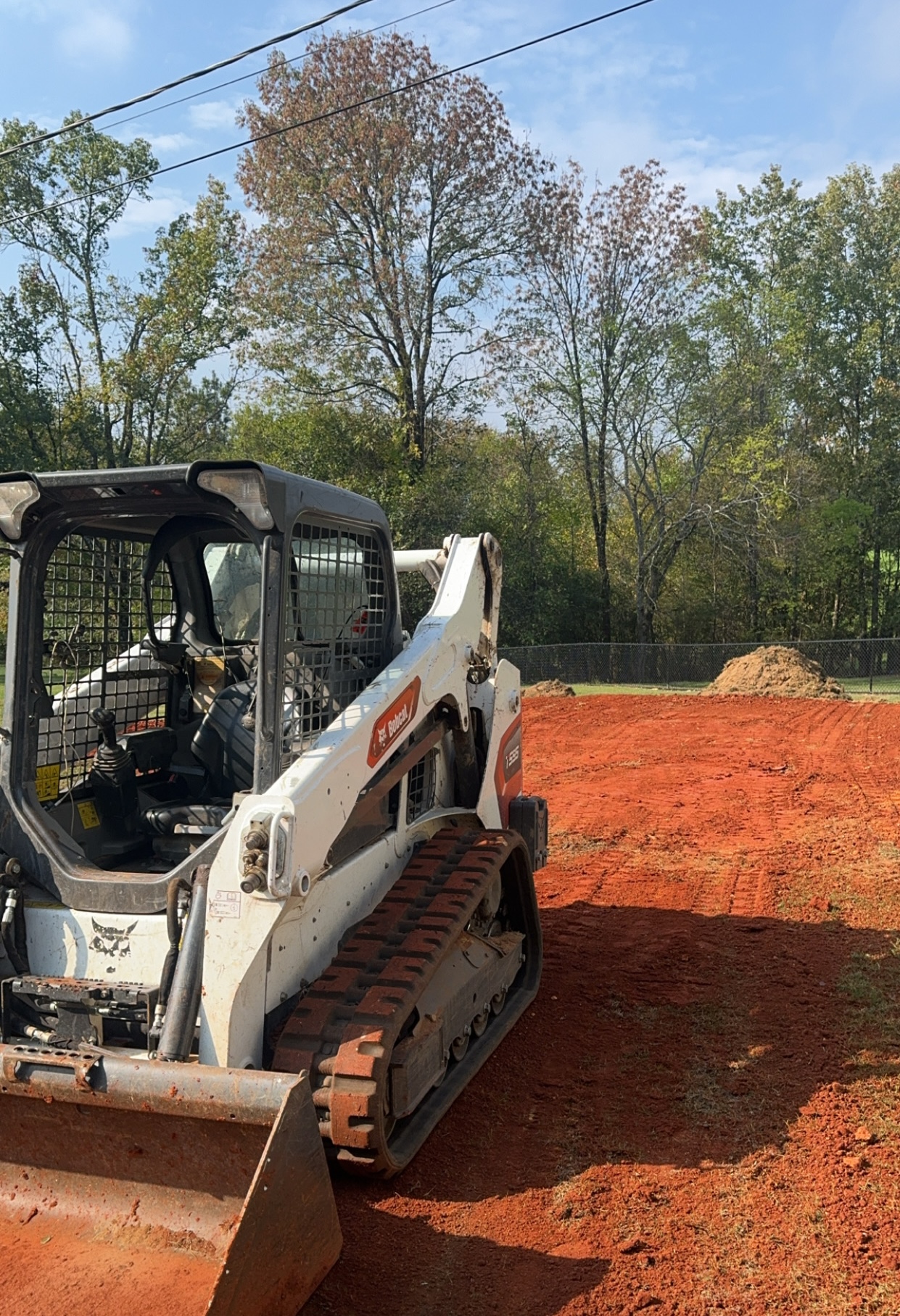 A compact track loader with a front bucket rests on red dirt, ideal for site preparation, with piles of soil and trees in the background on a sunny day.