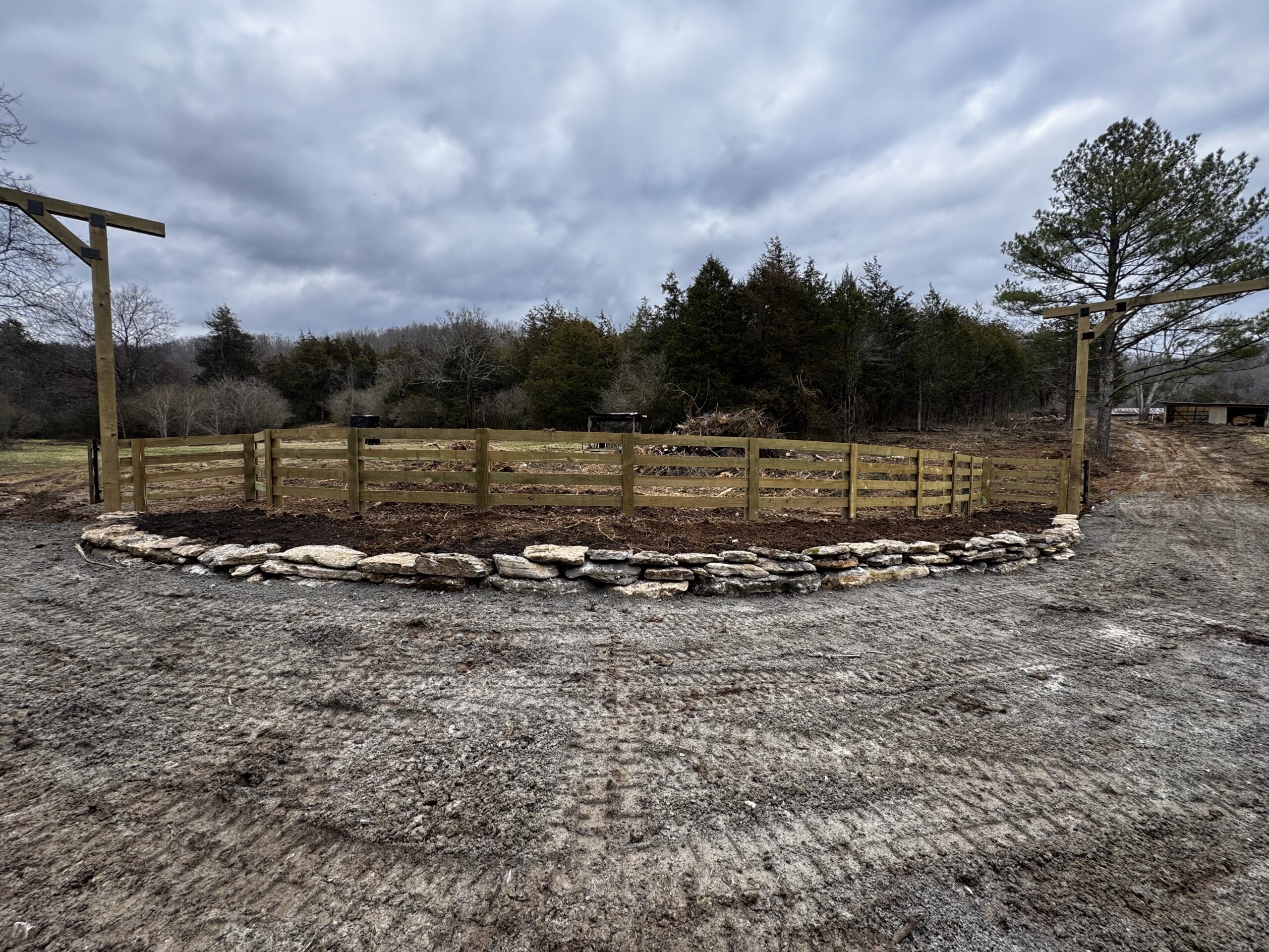 A circular wooden fenced area with a stone border sits on gravel, surrounded by trees under a cloudy sky—an ideal setting for slope stabilization or small retaining walls in your landscape.